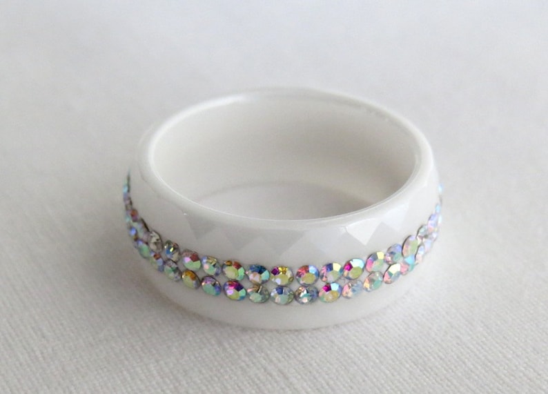 Puede incluir: Anillo de servilleta de cer&aacute;mica blanca con una banda de piedras de imitaci&oacute;n iridiscentes alrededor del borde superior.