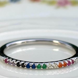 May include: A silver ring with a band of colorful gemstones. The gemstones are arranged in a rainbow pattern, including red, purple, black, blue, green, white, and orange. The ring is set on a white surface.