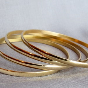 May include: Four gold-colored bangles are arranged on a light gray surface. The bangles are thin and have a smooth, shiny finish. The bangles are stacked together, creating a layered effect. The overall aesthetic is simple and elegant.