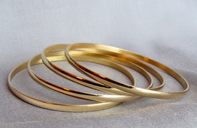May include: Four gold-colored bangles are arranged on a light gray surface. The bangles are thin and have a smooth, shiny finish. The bangles are stacked together, creating a layered effect. The overall aesthetic is simple and elegant.