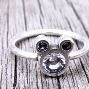 May include: A silver ring with a clear crystal in the center and two smaller black crystals on either side, shaped like Mickey Mouse's head.