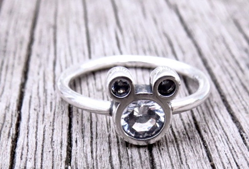 May include: A silver ring with a clear crystal in the center and two smaller black crystals on either side, shaped like Mickey Mouse's head.