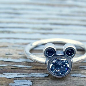 May include: A silver ring with a Mickey Mouse head design featuring a large clear crystal in the center and two smaller black crystals for the ears.