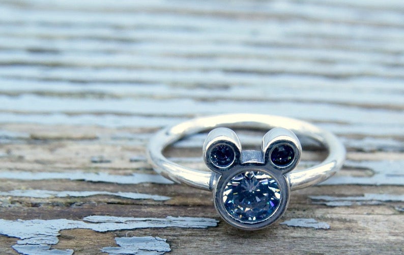 May include: A silver ring with a Mickey Mouse head design featuring a large clear crystal in the center and two smaller black crystals for the ears.