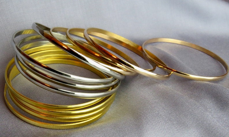 May include: A collection of gold and silver bangles. The bangles are stacked and arranged on a light gray, silky surface. The gold bangles have a bright, shiny finish, while the silver bangles have a polished, metallic appearance. The jewelry is simple and elegant.