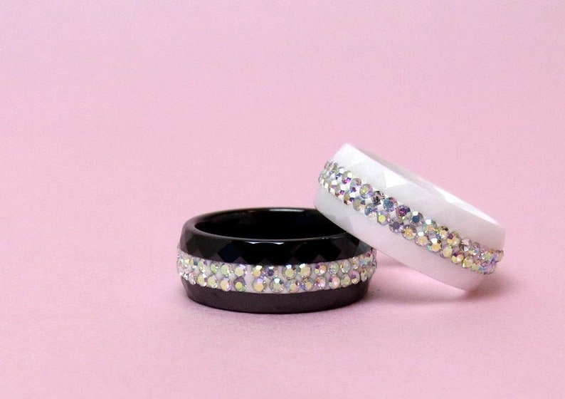 Puede incluir: Dos anillos con una fila de piedras de cristal iridiscentes. Un anillo es negro y el otro es blanco.