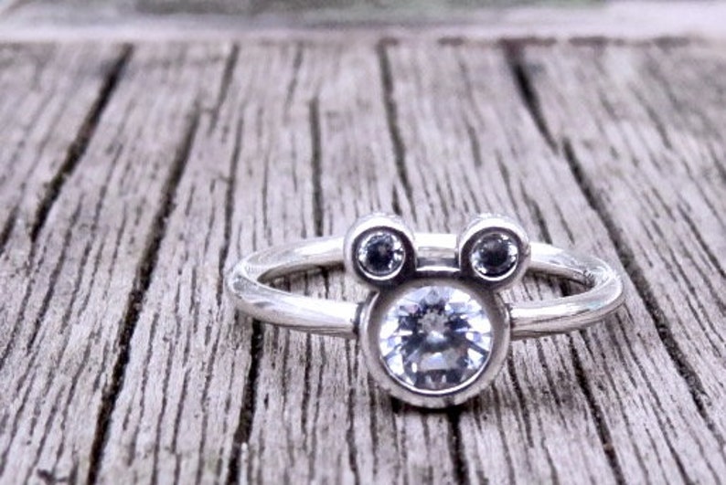 May include: A silver ring with a large clear stone in the center and two smaller clear stones on either side, shaped like Mickey Mouse ears.