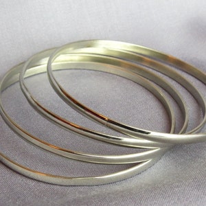 May include: Four silver bangles are arranged on a light purple fabric. The bangles are smooth, round, and reflective, with a simple, elegant design. The bracelets are a classic jewelry item.