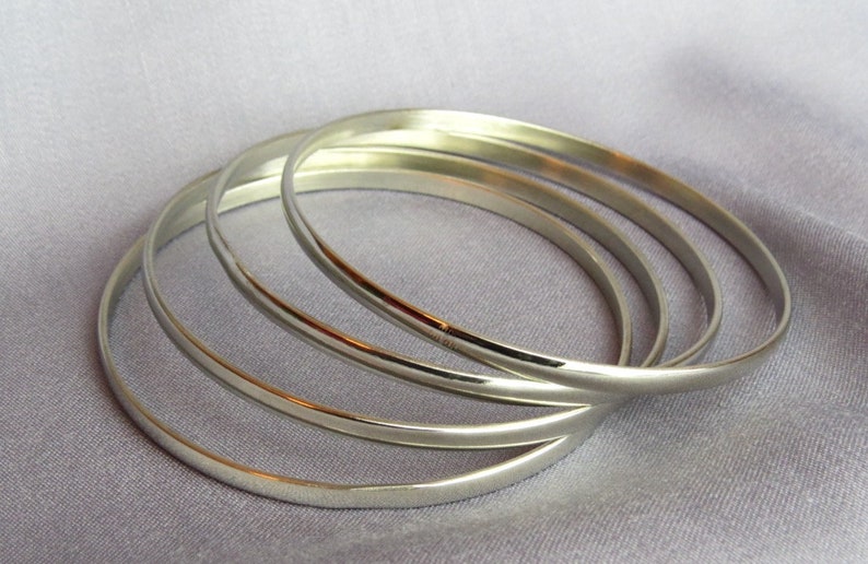 May include: Four silver bangles are arranged on a light purple fabric. The bangles are smooth, round, and reflective, with a simple, elegant design. The bracelets are a classic jewelry item.