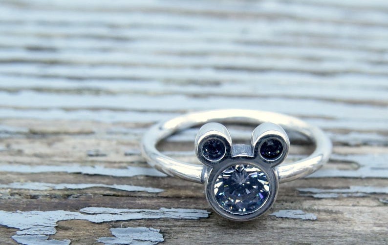 May include: A silver ring with a Mickey Mouse head design featuring a large clear rhinestone in the center and two smaller black rhinestones for the ears.
