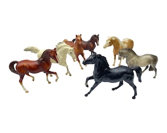 May include: A collection of seven miniature horse figurines in various colors, including brown, black, white, and gray. The figurines are posed in a standing position, with their heads turned in different directions.