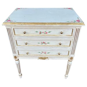 May include: A three-drawer chest with a light blue top and white body. The chest features gold trim, floral accents, and brass knobs. The legs are white with gold detailing, and the overall design is ornate.
