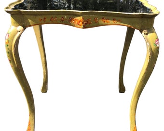 May include: A small, gold-painted table with a black glass top and floral details. The table has four legs with a curved design.