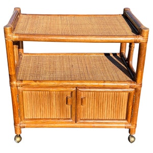 May include: A two-tiered bamboo serving cart with a woven rattan top and bottom shelves. The cart has two doors with a woven rattan design and rolls on four casters.
