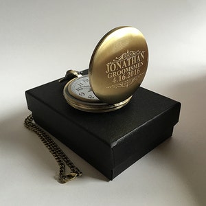 K&ouml;nnte beinhalten: Eine goldene Taschenuhr mit einer Kette in einer schwarzen Box. Das Zifferblatt der Uhr ist ge&ouml;ffnet und die R&uuml;ckseite ist mit dem Text "Jonathan Groomsmen 4.16.2016" graviert.