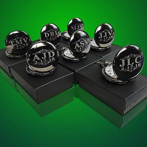 May include: A collection of eight silver pocket watches with engraved initials and dates. The watches are arranged on black boxes with a green background.