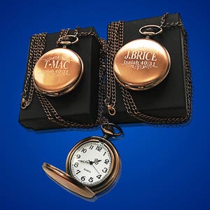 May include: Two gold pocket watches with chains on black boxes. One watch has the inscription "T-MAC Isaiah 40:31" and the other has the inscription "J.BRICE Isaiah 40:31".