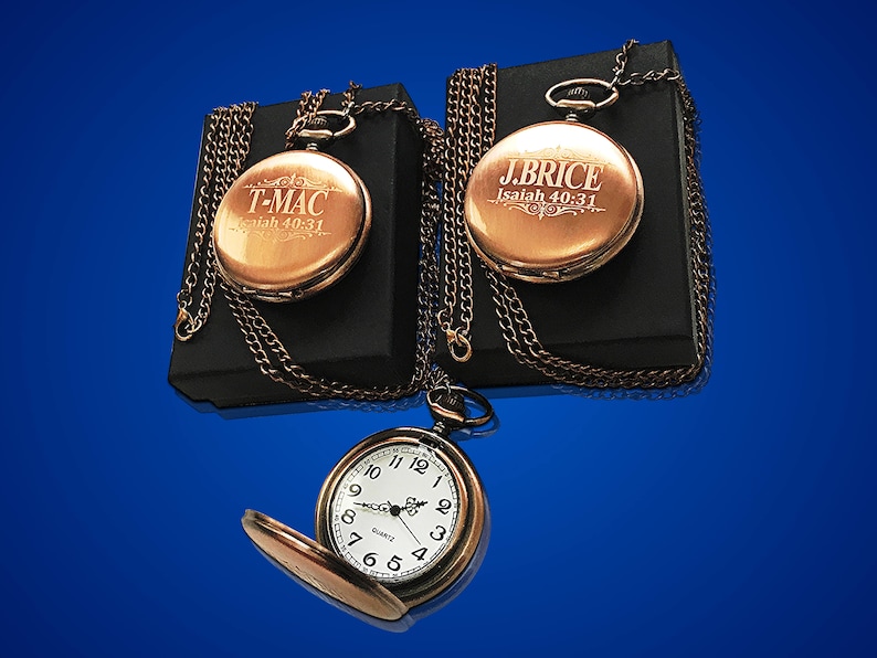 May include: Two gold pocket watches with chains on black boxes. One watch has the inscription "T-MAC Isaiah 40:31" and the other has the inscription "J.BRICE Isaiah 40:31".