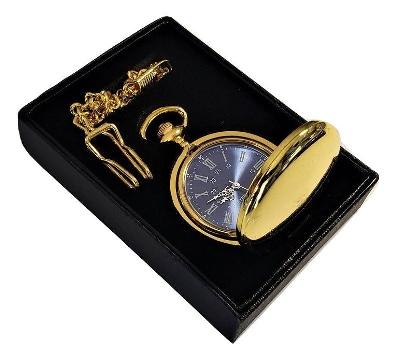 K&ouml;nnte beinhalten: Eine goldene Taschenuhr mit blauem Zifferblatt und r&ouml;mischen Ziffern f&uuml;r die Stunden. Die Uhr befindet sich in einer schwarzen Samtbox mit einer goldenen Kette.