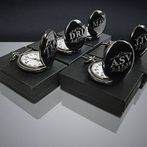 May include: Six black pocket watches with silver trim and engraved initials and dates in a silver font. The watches are displayed in black boxes.