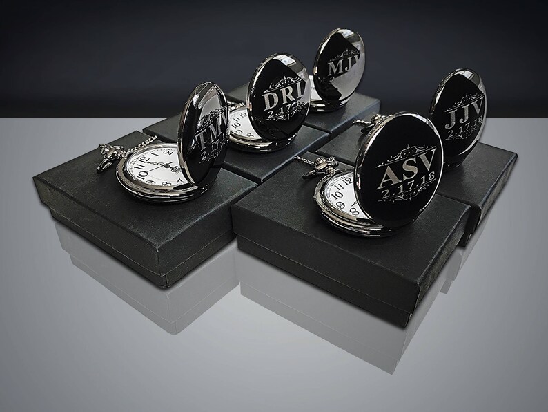 May include: Six black pocket watches with silver trim and engraved initials and dates in a silver font. The watches are displayed in black boxes.