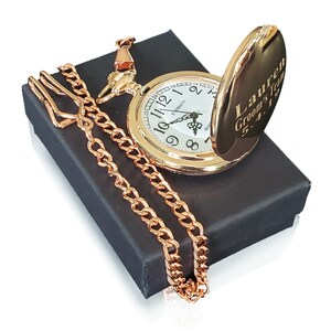 K&ouml;nnte beinhalten: Goldene Taschenuhr mit wei&szlig;em Zifferblatt und schwarzen r&ouml;mischen Ziffern. Die Uhr ist ge&ouml;ffnet und die R&uuml;ckseite ist mit dem Text "Lauren Groom's Team 5.4.19" graviert. Die Uhr wird mit einer goldenen Kette geliefert und befindet sich in einer schwarzen Box.