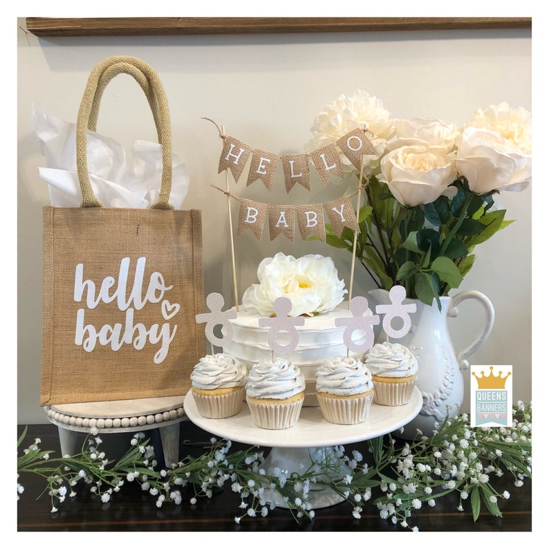 K&ouml;nnte beinhalten: Eine Jute-Tragetasche mit dem Text "hello baby" in wei&szlig;en Buchstaben. Die Tasche befindet sich neben einem wei&szlig;en Kuchen mit einem Jute-Banner, auf dem "hello baby" steht. Der Kuchen ist mit wei&szlig;em Frosting und rosa Schnuller-Toppern verziert. Es gibt auch Cupcakes mit rosa Schnuller-Toppern auf einem wei&szlig;en Kuchenst&auml;nder.