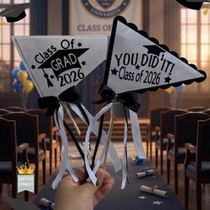 May include: Two silver graduation flag decorations with black trim and bows. One reads "Class Of Grad 2026" and the other says "You Did It! Class of 2026". The flags are on wooden sticks with white ribbons.