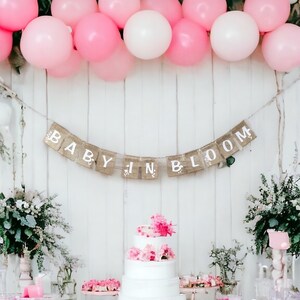 Op de afbeelding: Een roze en witte ballonversiering siert een witte muur met een banner waarop "Baby in Bloom" staat. Een witte vierlagentaart met roze bloemen staat op een tafel met roze en witte decoraties.