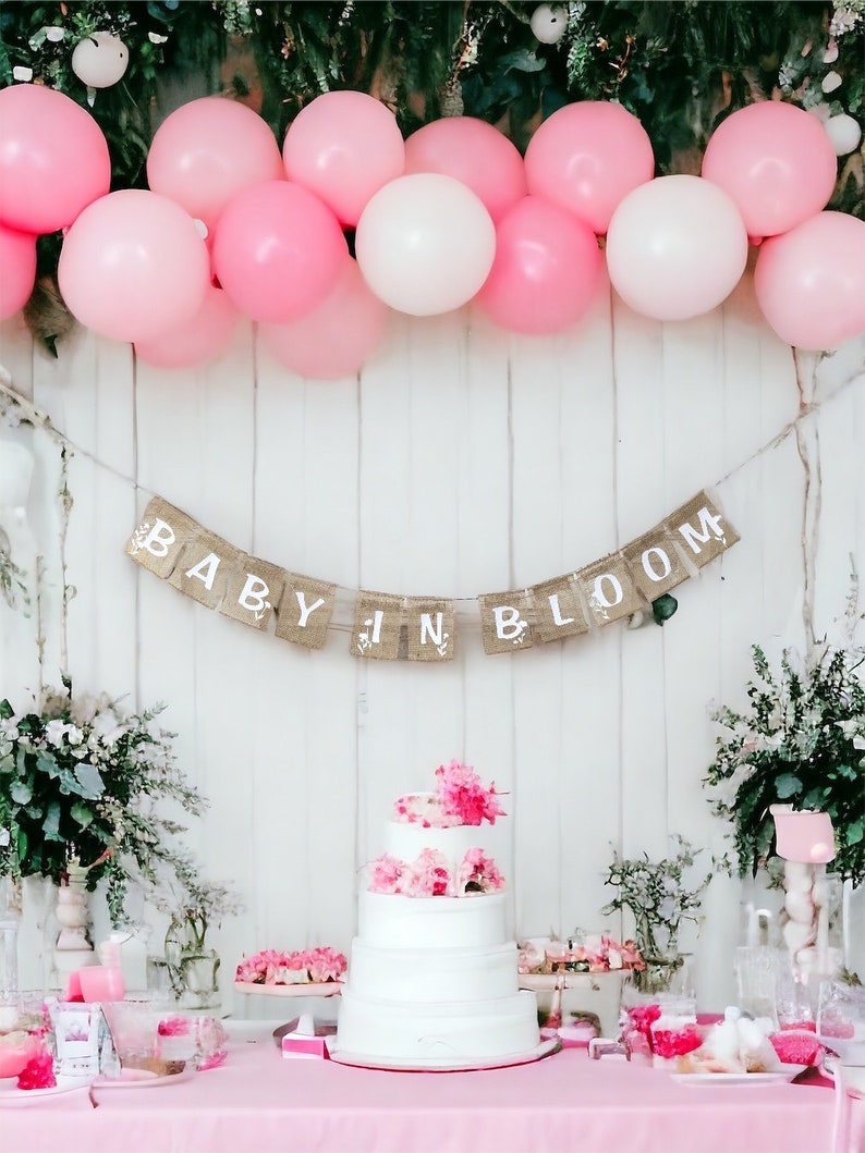 Op de afbeelding: Een roze en witte ballonversiering siert een witte muur met een banner waarop "Baby in Bloom" staat. Een witte vierlagentaart met roze bloemen staat op een tafel met roze en witte decoraties.