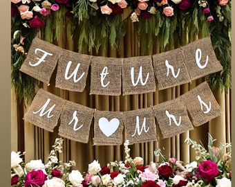 Könnte beinhalten: Ein Banner aus Jute mit den Worten "Future Mr & Mrs" in weißen Buchstaben. Das Banner ist mit einem weißen Herzen in der Mitte verziert. Das Banner hängt an einer Blumenkette.