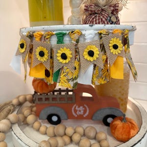 May include: A rustic fall-themed display featuring a miniature orange truck with the text "hello fall". The truck is surrounded by wooden beads, pumpkins, and a sunflower banner. A scarecrow doll and yellow jar complete the autumn arrangement.