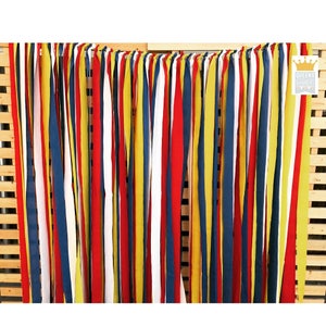 Puede incluir: Un tel&oacute;n de fondo colorido con tiras de tela verticales en rojo, amarillo, blanco y azul marino. Las tiras cuelgan de una estructura de madera, creando una exhibici&oacute;n vibrante y texturizada. El logotipo "QUEENS BANNERS" es visible en la esquina superior derecha.