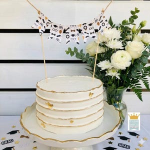 May include: A white frosted cake with gold trim and accents, topped with a banner featuring graduation-themed icons. The cake sits on a white pedestal with a gold rim, next to a vase of white flowers.