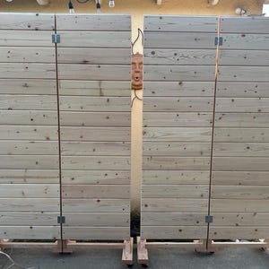 May include: Two wooden privacy screens with a light wood finish. Each screen is made of horizontal slats and has a hinged door. The screens are on wheels and can be moved around.