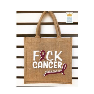 Op de afbeelding: Een bruine jute tas met witte tekst die "F*CK CANCER" zegt en een roze lint. De tekst "NEVER GIVE UP" staat onder het lint.