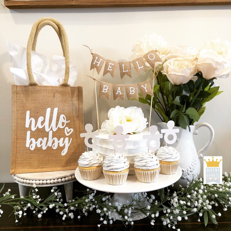 K&ouml;nnte beinhalten: Eine Jute-Tragetasche mit dem Text "hello baby" in Wei&szlig; mit einem kleinen Herz. Die Tasche steht auf einem wei&szlig;en Sockel. Es gibt einen Kuchen mit einem "hello baby" Banner darauf. Es gibt Cupcakes mit einem Babyrassel-Topper auf jedem Cupcake. Die Cupcakes stehen auf einem wei&szlig;en Kuchenst&auml;nder. Im Hintergrund befinden sich eine wei&szlig;e Kanne und ein Strau&szlig; wei&szlig;er Rosen.