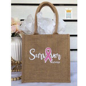 Op de afbeelding: Een bruine jute draagtas met een wit ontwerp dat "Survivor" zegt met een roze glitterlint en de tekst "Faith+Hope" eronder.