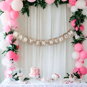 Op de afbeelding: Een baby shower decoratie met een roze en witte ballonboog, groen en een banner met de tekst "Baby in Bloom". Er is een tafel met een roze taart en andere desserts.
