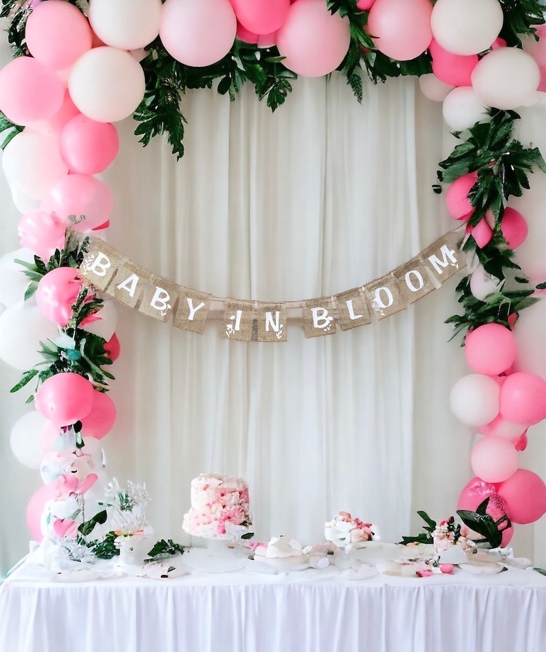 Op de afbeelding: Een baby shower decoratie met een roze en witte ballonboog, groen en een banner met de tekst "Baby in Bloom". Er is een tafel met een roze taart en andere desserts.