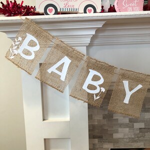 Op de afbeelding: Een banner van jute met het woord "BABY" gespeld in witte letters. Elke letter staat op een apart stuk jute en is versierd met kleine witte bloemen.