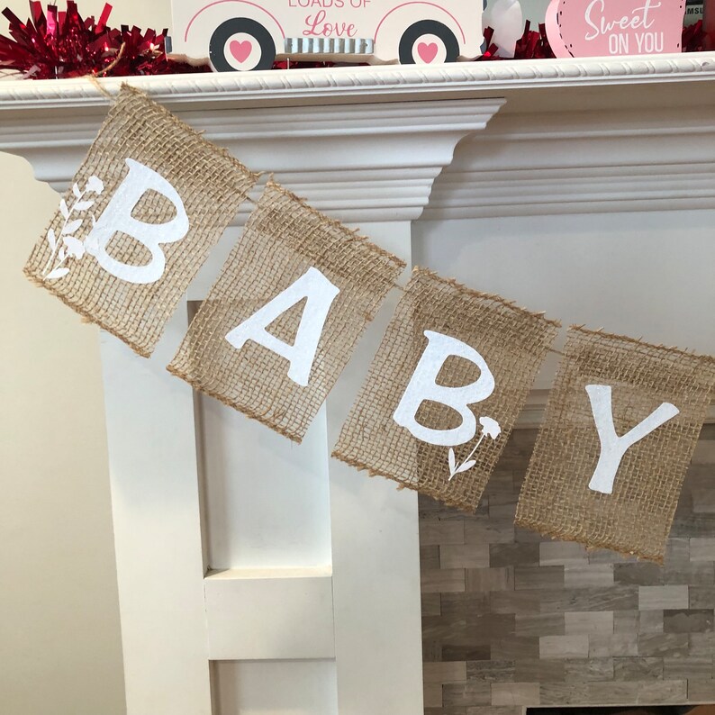 Op de afbeelding: Een banner van jute met het woord "BABY" gespeld in witte letters. Elke letter staat op een apart stuk jute en is versierd met kleine witte bloemen.