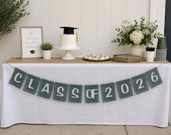 May include: A graduation party table setting with a white cake topped with a graduation cap, a banner that says "CLASS OF 2026", and a framed sign that reads "the TASSLE was worth the HASSLE". The table is decorated with flowers and snacks.