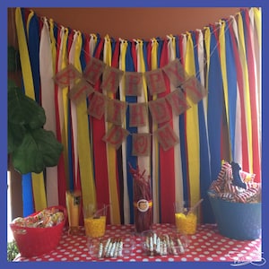 Puede incluir: Decoraci&oacute;n de fiesta de cumplea&ntilde;os con un mantel de lunares rojos y blancos. Una pancarta de arpillera con letras rojas dice "HAPPY BIRTHDAY". El fondo presenta cintas verticales de colores. Aperitivos y golosinas est&aacute;n dispuestos sobre la mesa.