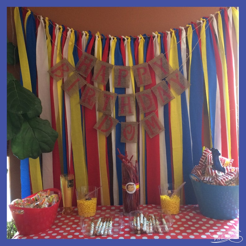 Puede incluir: Decoraci&oacute;n de fiesta de cumplea&ntilde;os con un mantel de lunares rojos y blancos. Una pancarta de arpillera con letras rojas dice "HAPPY BIRTHDAY". El fondo presenta cintas verticales de colores. Aperitivos y golosinas est&aacute;n dispuestos sobre la mesa.