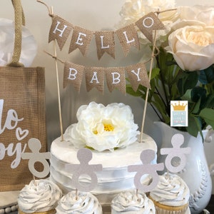 K&ouml;nnte beinhalten: Ein Banner aus Sackleinen mit dem Text "Hello Baby" darauf. Das Banner ist auf einem Holzstab befestigt und mit einer wei&szlig;en Blume verziert. Es gibt auch Cupcakes mit silbernen Glitzer-Schnuller-Toppern.