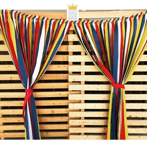 Puede incluir: Serpentinas de tela coloridas en rojo, amarillo, azul y blanco atadas con cintas rojas, colgando frente a un fondo de palets de madera. Las serpentinas forman parte de una exhibici&oacute;n decorativa, con el texto "QUEENS BANNERS" en la esquina superior derecha.
