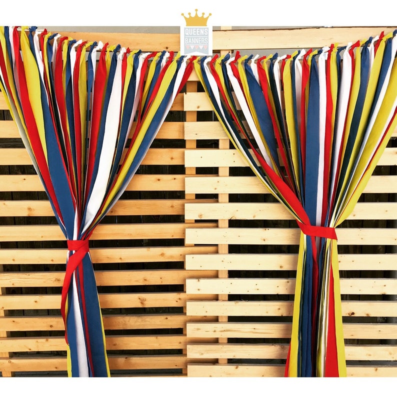 Puede incluir: Serpentinas de tela coloridas en rojo, amarillo, azul y blanco atadas con cintas rojas, colgando frente a un fondo de palets de madera. Las serpentinas forman parte de una exhibici&oacute;n decorativa, con el texto "QUEENS BANNERS" en la esquina superior derecha.