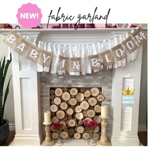 Op de afbeelding: Een banner van jute met witte stoffen kwastjes heeft de tekst "Baby in Bloom". De banner hangt boven een witte open haard met een houtkachel. De open haard is versierd met rode bloemen en kaarsen.