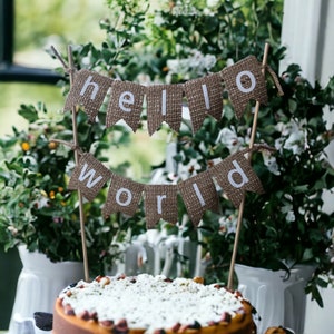 Op de afbeelding: Een versierde taart met een banner met de tekst "hello world". De banner is gemaakt van jute en heeft witte letters. De ronde taart heeft een witte topping met zichtbaar fruit en noten. Op de achtergrond witte vazen en groen.